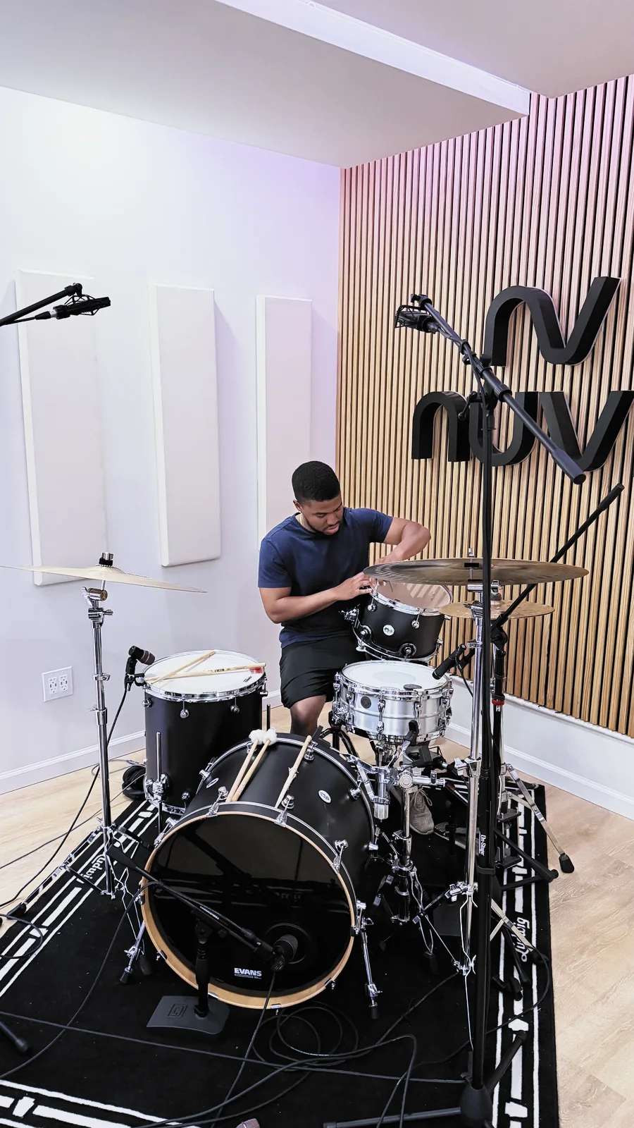 A drummer tracking in the Nuve live room — kit on a rug, overhead mics up, the backlit NUVE sign on the slatted wall behind.